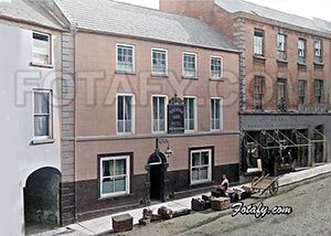 This is a remarkably restored 1900's image of the Beresford Hotel in Armagh. A man has unloaded a large quantity of luggage from a hand cart and sits on a wicker basket resting